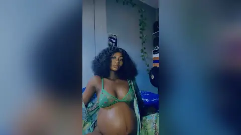 JoanJon is a pregnant woman with dark curly hair wearing a green lace bra while posing in her bedroom.