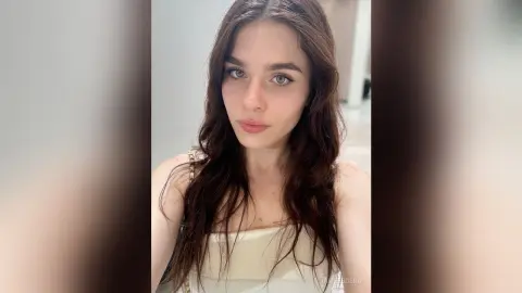 Young woman with long wavy brown hair wearing a white top in a bright, minimalist indoor setting.