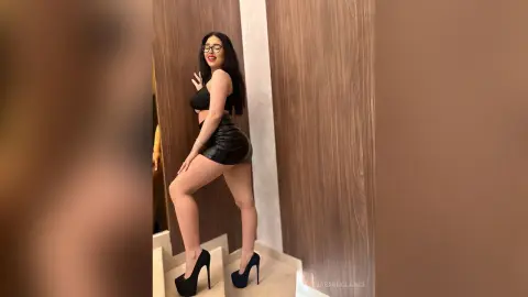 Smiling woman in a black crop top and leather mini skirt posing in a modern wood-paneled room.