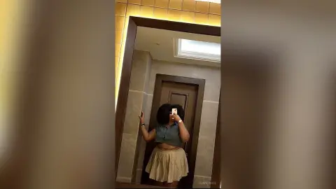 Young woman in a grey sleeveless top and beige skirt taking a mirror selfie in a modern hallway.