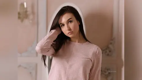 Young woman in a pink polka dot long-sleeve shirt posing with a soft, dreamy expression in a bright room.