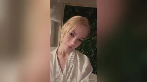 Blonde woman in a white waffle robe relaxing in a cozy, dimly lit room.