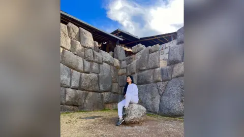 Young woman in white outfit sitting on stone ruins under a blue sky at an ancient Inca archaeological site.