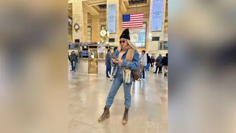 Stylish woman in denim jacket and beige scarf standing in a grand, sunlit historic train station lobby.