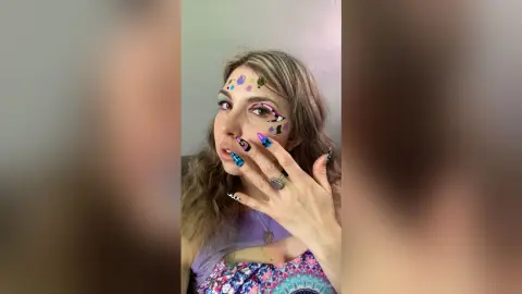Young woman with colorful face gems and long manicured nails posing in a casual purple top.