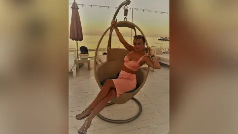 MilenaBia sits in a hanging egg chair on a deck wearing a peach-colored bra and skirt set.
