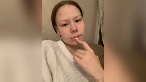 Young woman in a white ribbed sweater looking thoughtful in a cozy indoor setting.