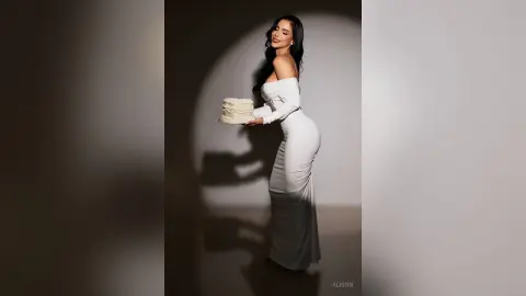 Alison poses in a white off-the-shoulder dress while holding a small white cake.