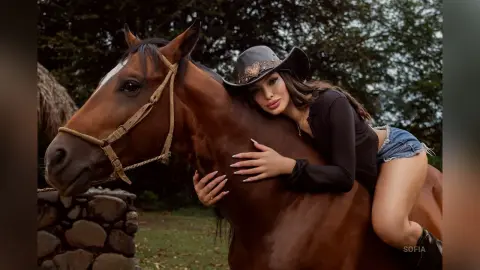 Stylish woman in black top and denim shorts leaning against a brown horse in a rustic outdoor setting.