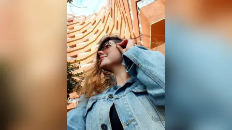 Smiling woman in a denim jacket and sunglasses posing outdoors against modern architecture.