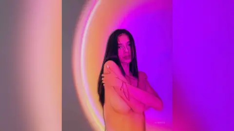 LucyMartin poses naked with her long dark hair, crossing her arms over her bare breasts against a purple and orange glowing background.