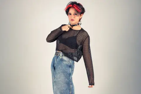 YuuMiko poses in a black mesh long-sleeve top, black bra, and denim skirt, wearing a black choker and showing her red-streaked hair against a plain background.
