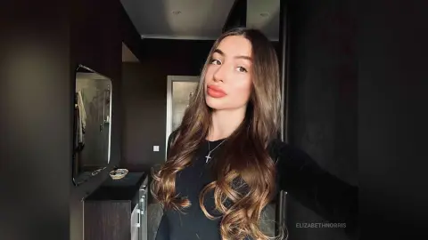 Young woman with long wavy hair wearing a black top and silver cross necklace in a dimly lit room.