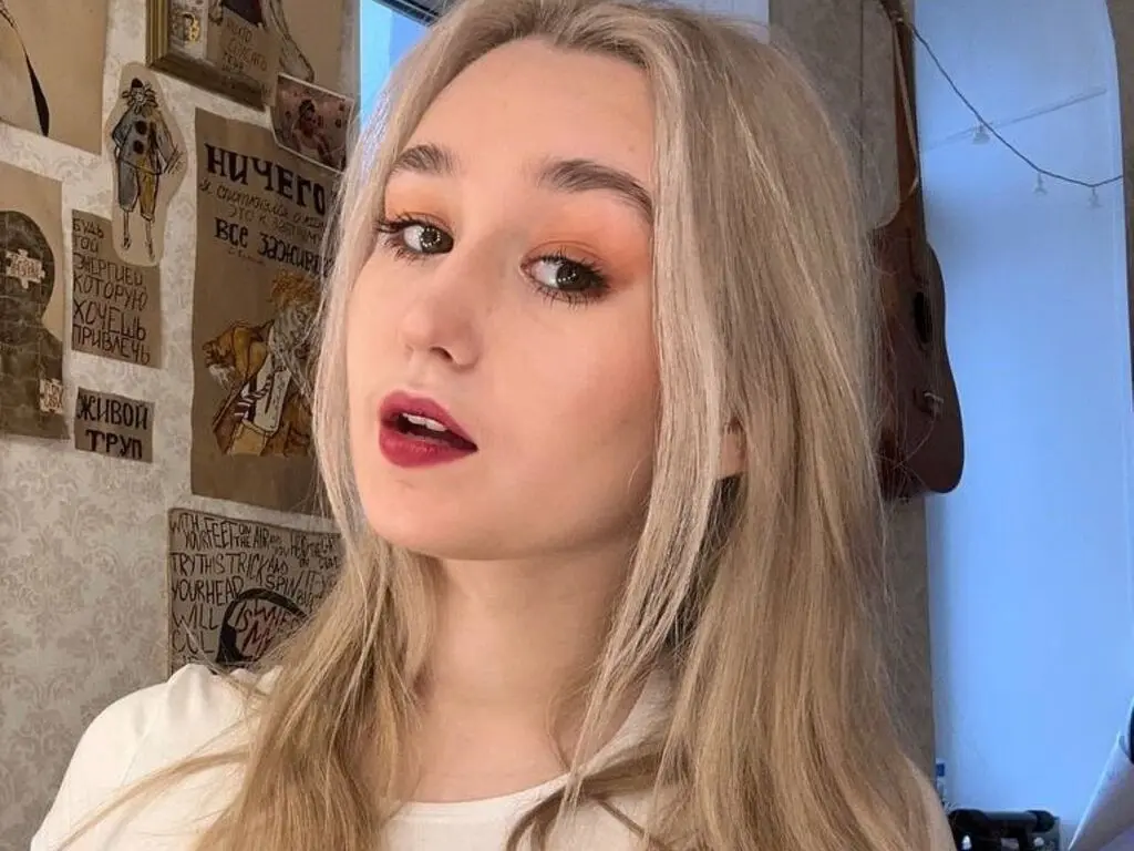 Blonde woman with warm makeup looking thoughtfully at the camera in a cozy room with vintage posters.
