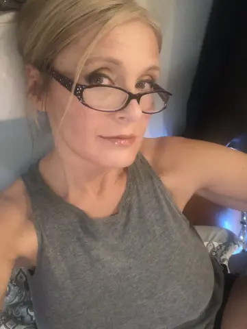 Blonde woman in a grey tank top wearing glasses, relaxing in a cozy indoor setting.