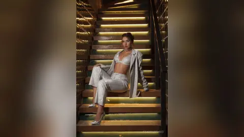 StacyHarland sits on a lit staircase wearing a white blazer and matching trousers with a white bra.