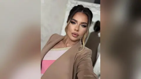 Glamorous woman in a beige blazer and pink striped top posing with elegant makeup in a bright room.