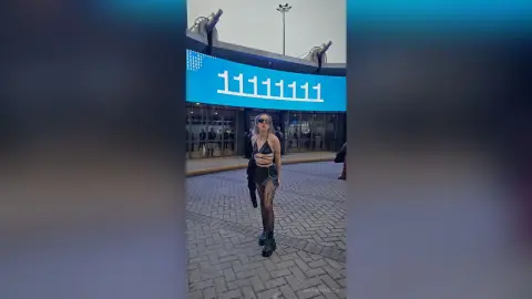 GiovannaKlein stands outdoors in a black cutout bodysuit and sheer tights, posing in front of a large blue digital sign.