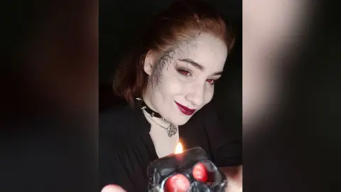 Gothic woman with red eyes and face vein makeup holding a skull candle in a dark, moody setting.