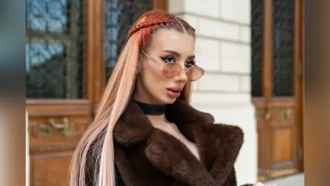 DeviNessa wears a black choker and a brown fur coat with her long, light-brown hair styled in a side braid.