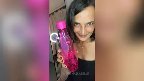 MichaelaBelle smiles while holding a pink bottle against her bare chest and neck.
