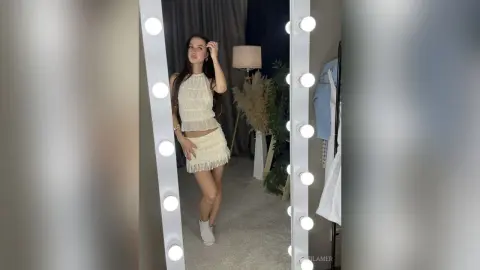 Young woman in a white lace halter top and mini skirt posing in a bright, modern bedroom mirror.