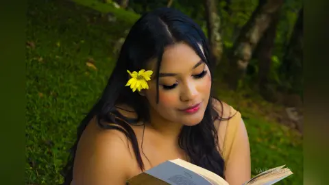 Young woman with a yellow flower in her hair reading a book in a lush green garden setting.