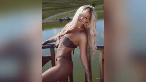 Anita poses on a wooden deck in a brown bikini, showing off her long blonde hair and tattoos.