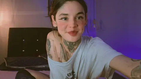 Smiling young woman with tattoos in a grey t-shirt relaxing in a dimly lit bedroom.