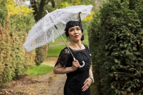 Elegant woman in a black lace dress holding a white lace parasol in a lush green garden setting.