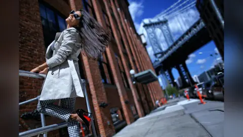 Stylish woman in a grey trench coat and checkered pants smiling on a sunny New York City street near a bridge.