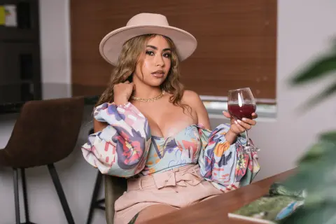 Stylish woman in a floral puff-sleeve top and wide-brimmed hat sipping red wine in a cozy indoor setting.