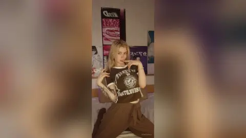 Blonde woman in a graphic t-shirt and brown trousers posing in a room with anime posters.