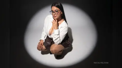 Elegant woman in a white button-down shirt and dark skirt posing under a dramatic spotlight in a studio.