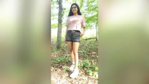 Young woman in a pink graphic t-shirt and black denim shorts standing in a lush green forest.