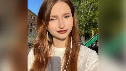 Young woman with long brown hair wearing a white ribbed top and wired earphones in a sunny outdoor park setting.
