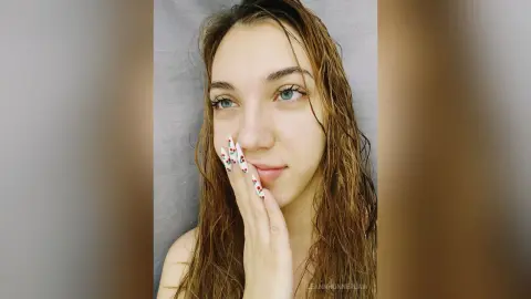 Young woman with wet hair and floral nail art looking thoughtfully at the camera in a soft indoor setting.