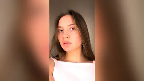 Young woman with long brown hair wearing a white tank top in a soft, minimalist studio setting.