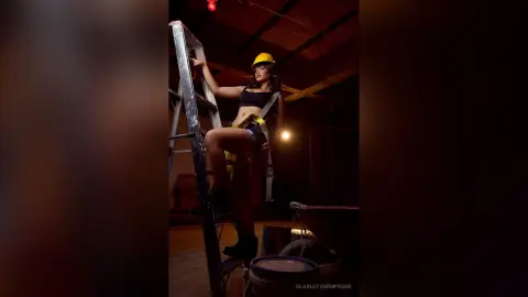 Young woman in black crop top and yellow hard hat posing on a ladder in a dark industrial warehouse setting.