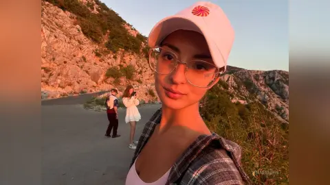 Young woman in glasses and white cap smiling during a sunset mountain hike in casual outdoor attire.