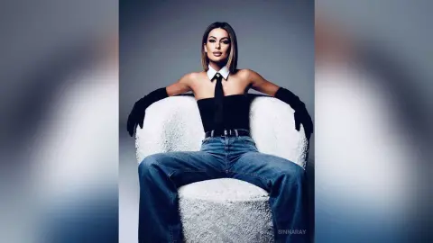 SinnaRay sits in a white chair wearing a black halter top, a black tie, and blue jeans with long black gloves.