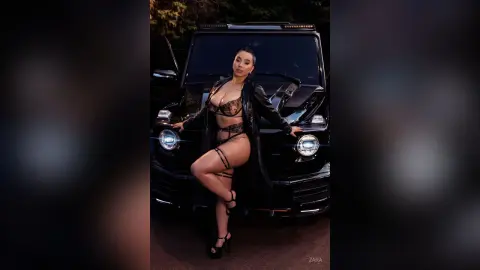 Confident woman in black lace lingerie and a leather jacket posing by a black SUV at dusk.