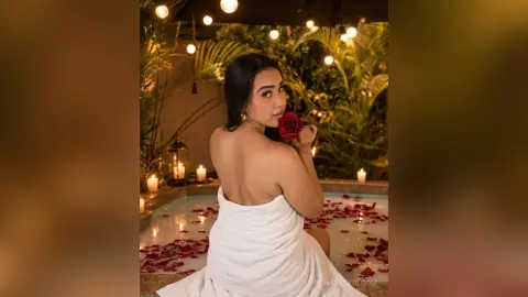 NatalyBloom sits in a lit pool surrounded by rose petals, looking over her shoulder while holding a red rose to her lips.