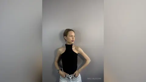 MalliaLucioni poses against a grey wall wearing a black halter top and light denim jeans, looking off to the side with her hands in her pockets.