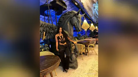 Stylish woman in a black bodysuit and trousers posing by a large grey horse statue in a moody, dimly lit lounge.