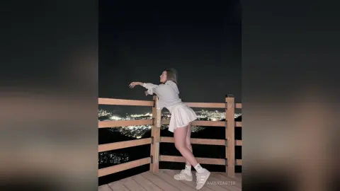 Young woman in a white long-sleeve top and mini skirt leaning on a balcony railing overlooking a city at night.