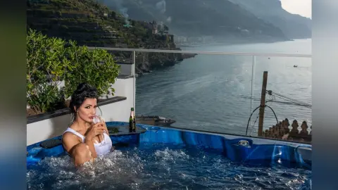 MissyJolie relaxes in a blue hot tub overlooking the ocean, wearing white lace lingerie while holding a glass of champagne.