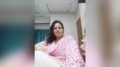 ManishaJi — ManishaJi smiles while lounging in a white and pink patterned saree in her room.