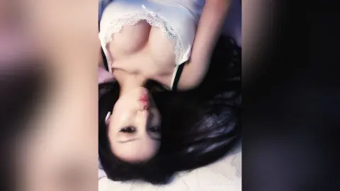 Young woman with long dark hair wearing a white lace camisole lying on a bed in a soft, moody atmosphere.