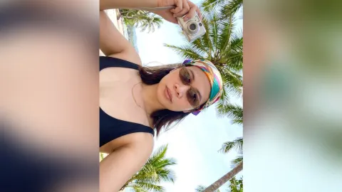 MilaSwan — MilaSwan wears a black bikini top and colorful headband while holding a silver camera under palm trees.